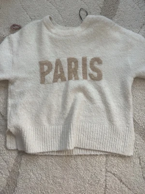 Vit stickad tröja med PARIS-tryck - Mysig vit stickad tröja från H&M Divided med beige texten 'PARIS' framtill. Tröjan har ribbstickad kant nedtill och en avslappnad passform. Perfekt för dig som gillar enkel men trendig stil.