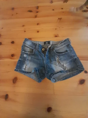 Blå jeansshorts från Bershka - Säljer ett par blå jeansshorts från Bershka med slitna detaljer och uppvikta benslut. Klassisk femficksmodell med knapp och dragkedja framtill. Perfekta för varma dagar och en avslappnad stil.