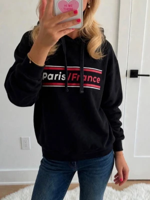  hoodie med Paris/France-tryck - Snygg  hoodie med huva och dragsnören. På bröstet står det 'Paris/France' i vitt och rött mot svarta och röda ränder. Hoodien har ribbade muddar vid ärmar och nederkant. Perfekt till jeans för en avslappnad look.