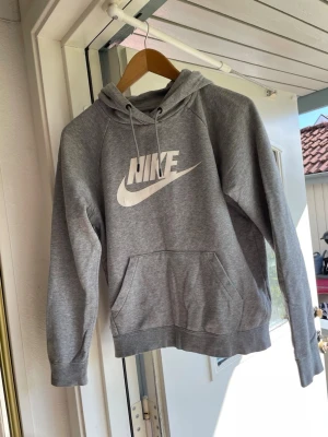 Grå hoodie från Nike med logga - Säljer en klassisk grå hoodie från Nike med stor vit logga på bröstet. Hoodien har huva med dragsko och en stor magficka. Tillverkad i mjukt sweatshirtmaterial och har ribbade muddar vid ärmar och nederkant. Perfekt för chill dagar eller till träningen. Obs logan är lite dålig så den har börjat lossna på vissa ställen.