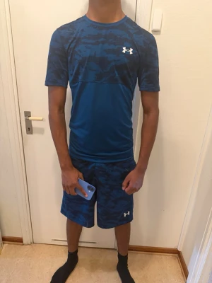 Blått träningsset från Under Armour - Säljer ett blått träningsset från Under Armour med t-shirt och shorts. Setet har ett snyggt kamouflagemönster upptill och på shortsen, samt logga på bröstet och benet. Tillverkat i ett lätt och snabbtorkande material som passar perfekt för träning.