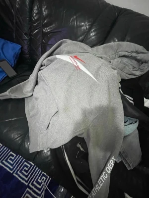 Grå Nike hoodie med röd logga - Säljer en grå hoodie från Nike med stor röd logga och texten 'NIKE ATHLETIC DEPT.' på bröstet. Hoodien har huva och är tillverkad i mjukt material, perfekt för en avslappnad och sportig look.