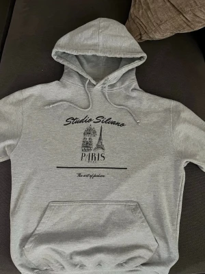 Studio Silano Paris Hodie - Snygg grå hoodie med tryck från Studio Silano. Framsidan har motiv av Eiffeltornet och texten 'Paris' samt 'The art of fashion'. Perfekt för en avslappnad streetwear-look. Hör gärna av dig vid funderingar