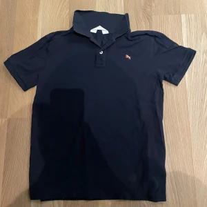 Mörkblå pikétröja - Säljer en mörkblå pikétröja från H&M med klassisk krage och två knappar framtill. Liten broderad logga i orange på bröstet. Tillverkad i 100% bomull och har korta ärmar. Perfekt för en stilren och enkel look.