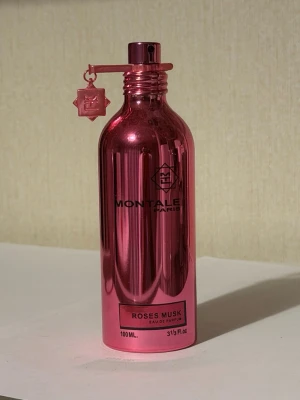 Montale Roses Musk Eau de Parfum - Montale Roses Musk Eau de Parfum i en metallicrosa flaska på 100 ml. Flaskan har en elegant, cylinderformad design med en dekorativ hängande charm vid sprayhuvudet. Doften kombinerar ros och mysk för en lyxig och modern känsla.