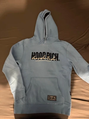  HOODRICH hoodie med tryck - Snygg ljusblå hoodie från HOODRICH med stor logga och grafiskt tryck både fram och bak. Klassisk känguruficka och justerbar huva. Tillverkad i mjukt bomullsmaterial för skön känsla. Perfekt för en avslappnad streetwear-look.