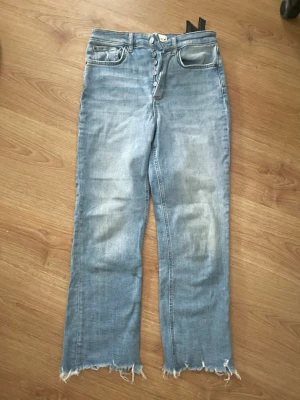 Ljusblå raka jeans med rå kant - Säljer ett par ljusblå jeans med raka ben och råa, fransiga benslut. Jeansen har knäppning framtill med flera knappar och klassiska fem fickor. Materialet är denim i bomull och de har en avslappnad passform.