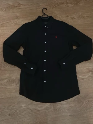 Svart skjorta från Polo Ralph Lauren - Svart skjorta från Polo Ralph Lauren med vit knappstängning och röd broderad logga på bröstet. Snygg för sommaren och hösten. Helt ny, använd en gång. Storlek M passar även S