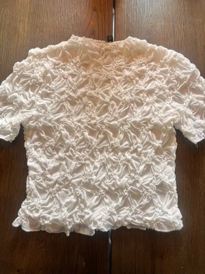 Vit volangblus från Vero Moda - Säljer en superfin vit blus från Vero Moda med korta ärmar och volangdetaljer över hela toppen. Materialet känns lätt och luftigt, perfekt för att lyfta vilken outfit som helst. Passformen är normal och toppen har en unik struktur som sticker ut.