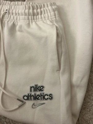 Vita mjukisbyxor från Nike Athletics - Säljer ett par vita mjukisbyxor från Nike Athletics med broderad logga och dragsko i midjan. Byxorna har resår i midjan och sidofickor. 