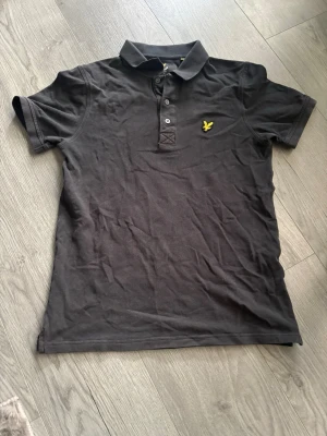 Svart pikétröja från Lyle & Scott  - Tjena! Jag säljer en svart pikétröja i bomull med klassisk krage och knappar framtill. Tröjan har en gul broderad logga på bröstet och korta ärmar. Perfekt till en avslappnad stil. Den är i ganska bra skick men lite lite sliten.