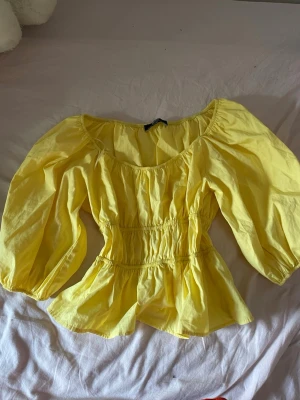 Gul blus med puffärmar från Bershka - Söt gul blus från Bershka med puffiga långärmar och smockad midja som ger en snygg siluett. Blusen har en rundad halsringning och volang nedtill. Perfekt för dig som vill ha en färgglad och trendig topp till sommaren.