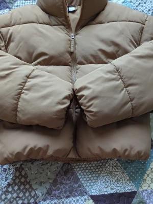 Brun pufferjacka från H&M - Säljer en brun pufferjacka med hög krage och dragkedja framtill. Jackan har en boxig passform och breda, vadderade paneler som ger en riktigt puffig look. Perfekt för kalla dagar när du vill hålla dig varm och stilsäker. Storleken passar för XS också. 
