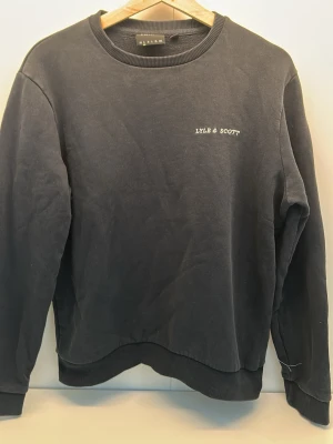 Svart sweatshirt från Lyle & Scott - Klassisk svart sweatshirt från Lyle & Scott med rund halsringning och diskret broderad logga på bröstet. Tröjan har ribbade muddar vid ärmslut och nederkant. Perfekt för en avslappnad och stilren look.