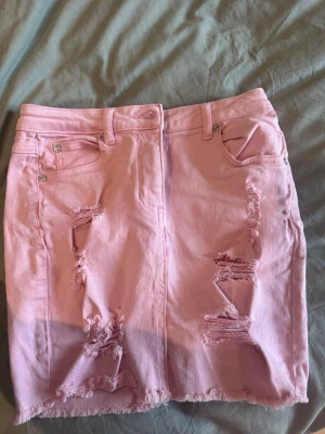 Rosa kort jeanskjol med slitningar - Säljer en ljusrosa kort jeanskjol med slitna detaljer och rå kant nertill. Kjolen har femficksmodell, bälteshällor och dragkedja med knapp framtill. Perfekt för dig som vill ha en edgy och trendig look.