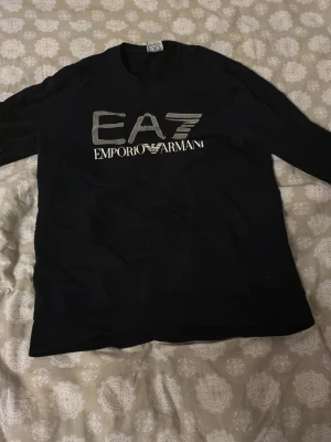 Svart tröja från Emporio Armani EA7 - Snygg svart långärmad tröja från Emporio Armani EA7 med stor logga framtill i vitt och silver. Klassisk rund halsringning och mjukt material som känns skönt mot huden. Perfekt för dig som gillar stilrena och exklusiva plagg.