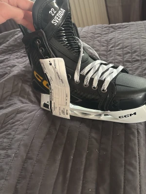 CCM FT2 hockeyskridskor svart/vit - Säljer ett par grymma CCM FT2 hockeyskridskor i svart med vita och gula detaljer. Skridskorna har klassisk snörning, robust kolfiberdesign och vita skenor. Perfekta för dig som vill ta din hockey till nästa nivå.