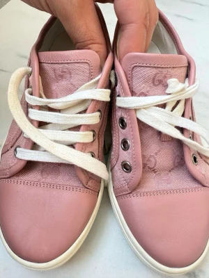 Rosa sneakers från Gucci - Säljer ett par snygga rosa sneakers från Gucci med vita snören och vita sulor. Skorna har klassisk låg modell och är tillverkade i en mix av läder och textil med diskret GG-mönster på sidan och plös. Perfekta för dig som gillar exklusiv streetstyle.