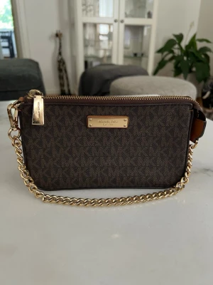 Brun väska Michael Kors - Snygg brun kuvertväska från Michael Kors med guldiga detaljer och kedja. Väskan har MK-mönster över hela ytan och en metallbricka framtill. Praktisk dragkedja upptill och stilren design i tåligt material.