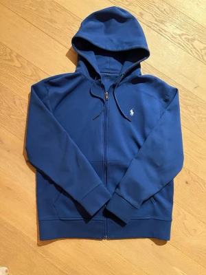 Ralph Lauren Hoodie. Helt ny - Helt ny snygg blå hoodie från Polo Ralph Lauren med dragkedja framtill och klassisk gul broderad logga på bröstet. Hoodien har huva med dragsko, två fickor och ribbade muddar. Perfekt för en chill och stilren look.