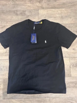 Svart t-shirt från Polo Ralph Lauren - Klassisk svart t-shirt från Polo Ralph Lauren med liten vit logga broderad på bröstet. T-shirten har rund hals och korta ärmar, perfekt till jeans eller shorts. Tillverkad i mjuk bomull för skön känsla.