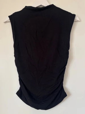 Svart ärmlös topp med rynkade sidor - Säljer en svart ärmlös topp med rynkade detaljer längs sidorna. Toppen har en enkel och clean design med rund hals och är tillverkad i mjukt bomullstyg. Perfekt att styla med jeans eller kjol för en chill look.