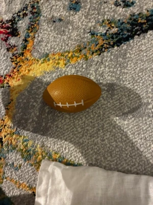 Liten brun amerikansk fotboll - En liten amerikansk fotboll i brun färg med vit söm. Bollen är oval och har en texturerad yta som ger bra grepp. Perfekt för lek eller dekoration. Är även en squishy.