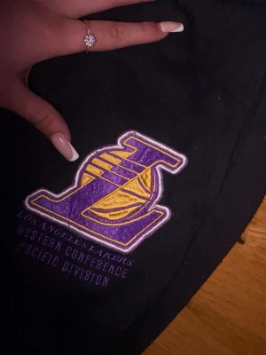 Svarta Lakers mjukisbyxor - Säljer ett par svarta mjukisbyxor med Los Angeles Lakers-logga i lila och gult på benet. Byxorna har avslappnad passform och resår i midjan. Perfekta för dig som gillar basket och vill ha en sportig stil.