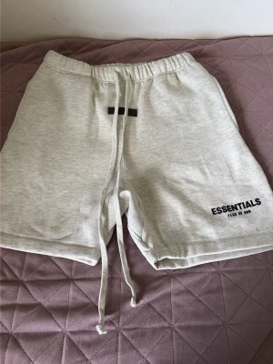 Grå shorts från Essentials Fear of God - Snygga ljusgrå shorts från Essentials Fear of God med långa snören i midjan och tryckt logga på benet. Mjukt material och relaxed fit, perfekta för chill dagar eller streetwear-stil.