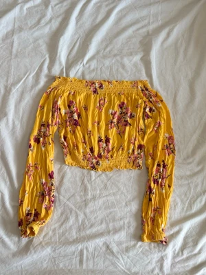 Gul offshoulder blommig topp - Säljer en gul offshoulder topp med blommigt mönster i rosa och lila toner. Toppen har långa ärmar med resår vid handleder och midja för en snygg croppad look. Perfekt för sommarens alla tillfällen!