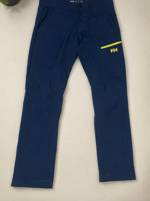 Blå byxor från Helly Hansen - Mörkblå byxor från Helly Hansen med raka ben och gul dragkedja på benfickan. Byxorna har en sportig look och är tillverkade i ett syntetmaterial som känns lätt och smidigt. Perfekta för dig som gillar funktionella och stilrena plagg.