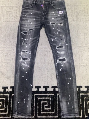 Grå slitna skinny jeans med färgstänk från disquared  - Säljer ett par grå skinny jeans med slitningar och hål på framsidan samt vita färgstänk över hela byxan. Jeansen har fem fickor och en patchdetalj vid fickan fram. Perfekta för en edgy och trendig streetwear-look.