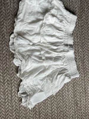 Vit kjol med volang och snörning - Säljer ett par vita shorts med volangkant och snörning framtill. De har elastisk midja och är tillverkade i ett lätt, luftigt material som passar perfekt till varma dagar. Stilen är loose och de har en feminin touch med de fina detaljerna.