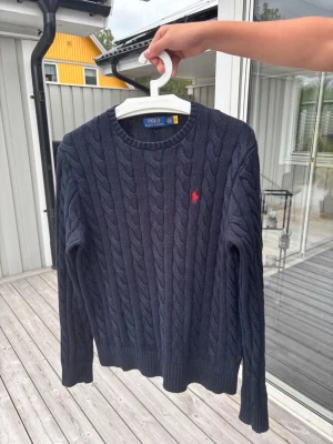 Mörkblå kabelstickad tröja Polo Ralph Lauren - Mörkblå kabelstickad tröja från Polo Ralph Lauren med rund halsringning och röd broderad logga på bröstet. Tröjan har långa ärmar och ribbade muddar vid ärmslut och nederkant. Perfekt för en stilren och klassisk look.