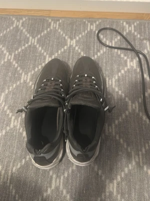 Svarta sneakers med vita detaljer - Säljer ett par svarta sneakers med vita detaljer och snörning. Skorna har en sportig design med mesh- och syntetmaterial samt en tjock vit sula. Perfekta för dig som gillar en clean och stilren look till vardags.