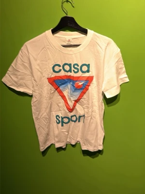 Vit Casa Sport t-shirt med tryck - Vit t-shirt med färgglatt Casa Sport-tryck framtill i blått och rött. Klassisk rund hals och korta ärmar. Tillverkad i mjuk bomull, perfekt för en avslappnad och sportig stil.