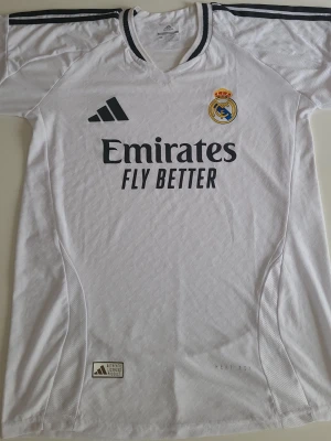 Real Madrid matchtröja Adidas vit - Snygg vit matchtröja från Real Madrid med klubbmärke på bröstet och Adidas-logga. Tröjan har svarta detaljer på axlarna och sponsortryck framtill. Tillverkad i lätt och ventilerande funktionsmaterial, perfekt för fotboll eller träning.