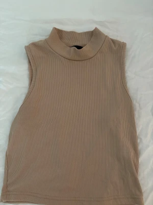 Beige ribbad ärmlös polotopp - Säljer en beige ärmlös polotopp med ribbad struktur. Toppen har hög krage och är i ett mjukt, stretchigt material som sitter snyggt på kroppen. Perfekt att styla med jeans eller kjol för en trendig look.
