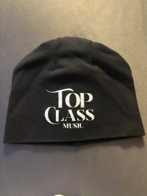 Svart mössa med Top Class Music-tryck - Snygg svart mössa med vit text där det står 'Top Class Music' framtill. Tillverkad i mjukt och stretchigt tyg som sitter skönt på huvudet. Perfekt accessoar för dig som gillar musik och vill ha en stilren look.