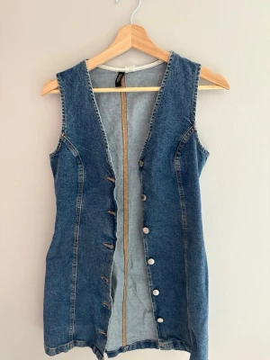 Ärmlös jeansklänning med knappar - Snygg ärmlös klänning i blå denim med knappar framtill. Klänningen har en figurnära passform och klassiska jeanssömmar. Använd en gång. Skriv privat för mer information. 