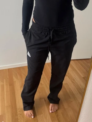 Svarta överdragsbyxor från Adidas - Säljer ett par svarta träningsbyxor från Adidas med dragsko i midjan och raka ben. Byxorna har en vit Adidas-logga på vänster lår.