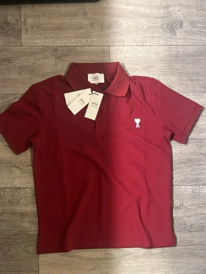 Ami polo shirt - Snygg röd pikétröja från Ami Paris med klassisk krage och korta ärmar. Tröjan har en vit broderad logga med hjärta och bokstaven A på bröstet. Perfekt för en stilren och avslappnad look.