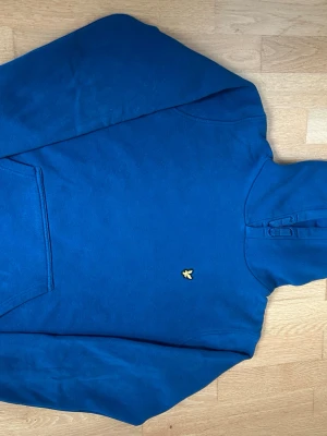 Blå hoodie Lyle and Scott - Snygg blå hoodie med huva och magficka. På bröstet sitter en liten gul broderad logga som ger en clean och stilren look. Hoodien har ribbade muddar vid ärmslut och nederkant. Perfekt för dig som vill ha en enkel men ändå trendig stil.