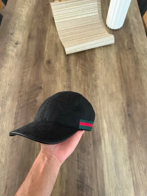 Gucci keps  - Snygg svart keps med diskret mönster och en grön-röd rand på sidan. Klassisk böjd skärm och justerbar passform. Materialet ser ut att vara tyg med en exklusiv känsla. Perfekt för dig som vill ha en stilren accessoar med en touch av färg.