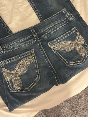 Mörkblå jeans med broderade bakfickor! - Säljer ett par mörkblå lågmidjade jeans från GinaTricot med snygga broderade detaljer och paljetter på bakfickorna. Jeansen har kontrastsömmar och klassisk femficksmodell. Materialet är denim i bomull med lite stretch för extra komfort. Jeansen är helt nya men utan prislapp💕