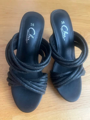 Svarta pumps med flätade remmar - Snygga svarta pumps från Clou i storlek 36. Skorna har höga klackar och flera flätade remmar över foten som ger en cool och trendig look. Tillverkade i syntetmaterial med öppen tå och slip-in modell.