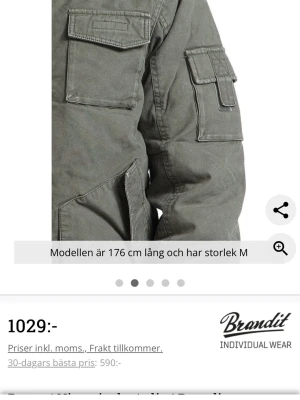 Grön parkasjacka från Brandit - Stilren grön parkasjacka från Brandit med flera praktiska fickor på bröstet och ärmen. Jackan har huva och detaljerade sömmar för en cool look. Perfekt för dig som vill ha en funktionell och snygg jacka till höst och vinter.