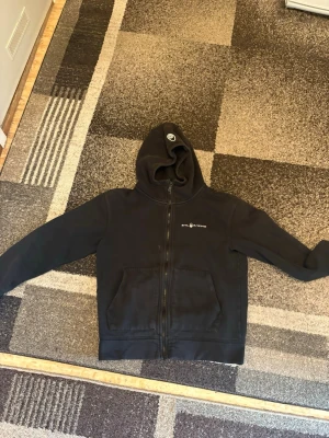 Svart hoodie med dragkedja - En svart hoodie med dragkedja framtill och två stora fickor. Tröjan har huva med snörning och diskret tryck på bröstet. Perfekt för chill dagar eller när du vill ha något bekvämt och enkelt. Klassisk passform och mjukt material.