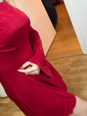 Röd sammetsklänning med lång ärm - Snygg röd klänning i mjuk sammet med lång ärm och lätt utsvängda ärmslut. Klänningen har en figurnära överdel och faller fint från midjan. Perfekt för dig som vill sticka ut med en elegant look.