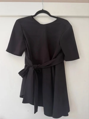 Svart blus från Zara Woman - Stilren svart blus från Zara Woman med knytband i midjan och korta ärmar. Tröjan har en enkel, elegant design och rund halsringning. Perfekt för dig som gillar minimalistisk stil.